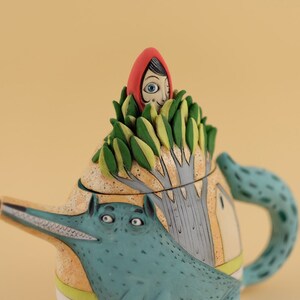 May include: A ceramic teapot shaped like a wolf with a green leafy tree on top. A girl wearing a red hood is peeking out from the top of the tree.