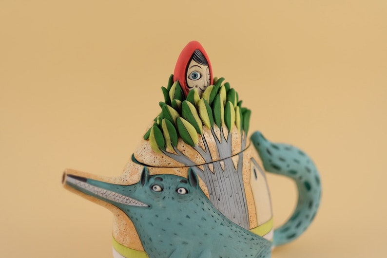 May include: A ceramic teapot shaped like a wolf with a green leafy tree on top. A girl wearing a red hood is peeking out from the top of the tree.