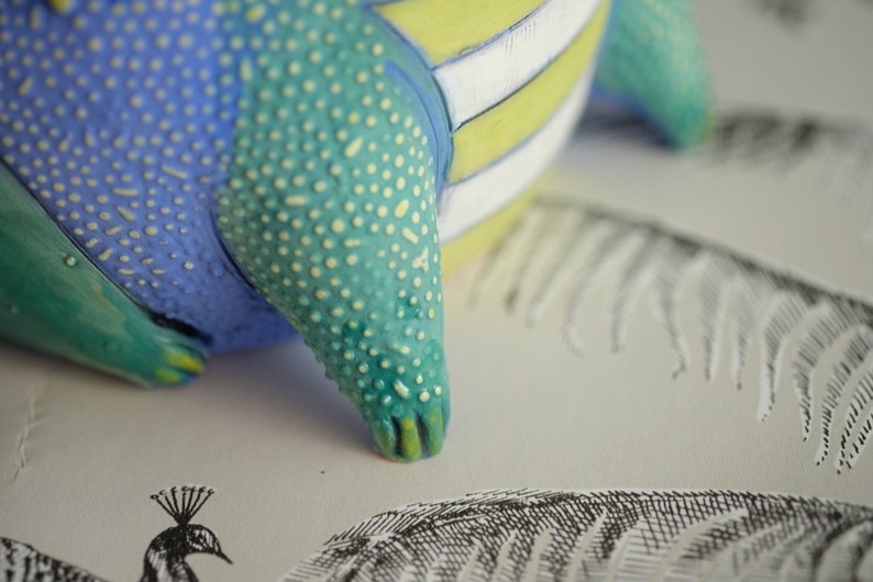 May include: A ceramic figurine with a blue, green, and yellow striped body. The figurine has a textured surface and a pointed tail.
