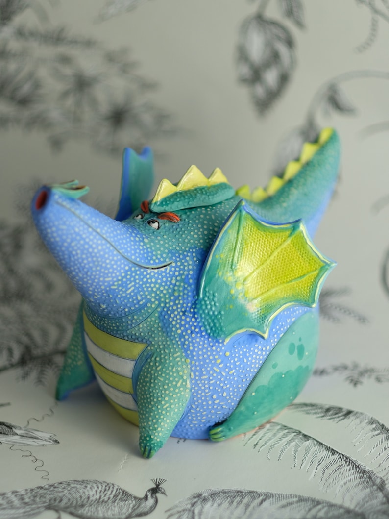 May include: A blue and green ceramic dragon teapot with yellow wings and a white stripe around its belly. The teapot has a whimsical, cartoonish design.