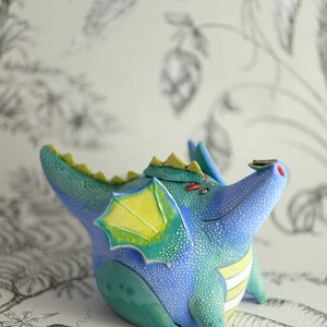 May include: A blue and green ceramic dragon teapot with yellow wings and white stripes. The dragon is sitting on a white surface with a black and white floral pattern.