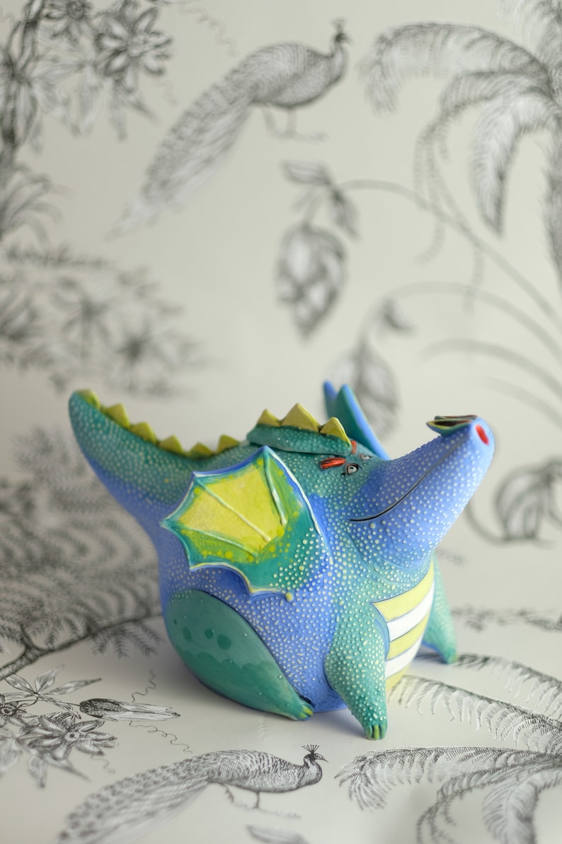 May include: A blue and green ceramic dragon teapot with yellow wings and white stripes. The dragon is sitting on a white surface with a black and white floral pattern.