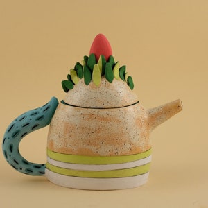 May include: A whimsical ceramic teapot with a speckled orange glaze, green and yellow leaf accents, and a bright red top. The teapot has a blue and white patterned handle.