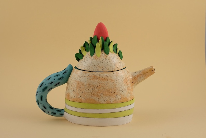 May include: A whimsical ceramic teapot with a speckled orange glaze, green and yellow leaf accents, and a bright red top. The teapot has a blue and white patterned handle.