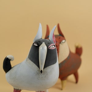 May include: Two quirky ceramic sculptures. The front figure is light grey with a black head and beak, pink feet, and a black and white tail. The second figure is reddish-brown with white and pink accents. Both have exaggerated features.