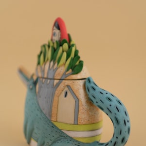May include: A ceramic teapot shaped like a wolf with a house on its back. The house has a red roof and a green tree growing out of it. The teapot is painted in shades of blue, green, yellow, and white.