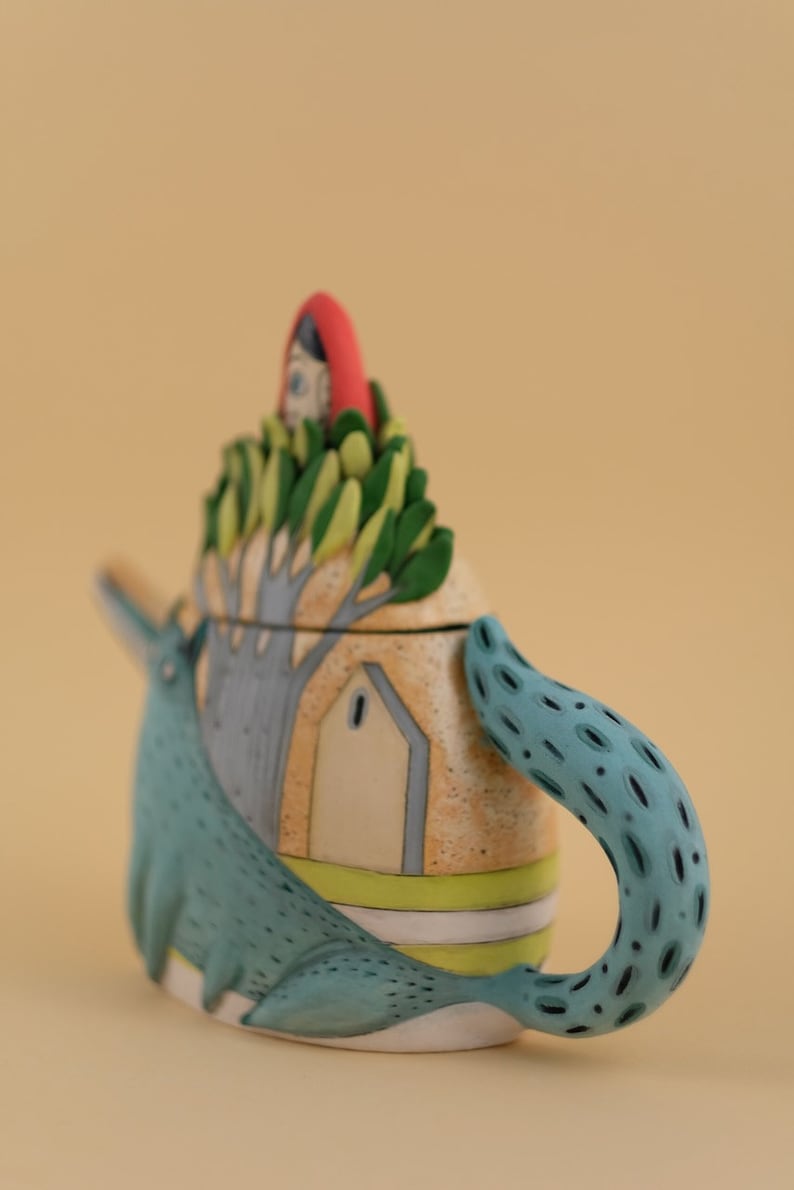 May include: A ceramic teapot shaped like a wolf with a house on its back. The house has a red roof and a green tree growing out of it. The teapot is painted in shades of blue, green, yellow, and white.