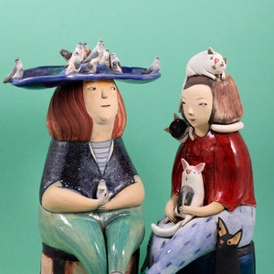 Może przedstawiać: Dwie ceramiczne figurki kobiet, każda z innym wyrażeniem twarzy. Kobieta po lewej stronie nosi niebieski kapelusz z ptakami i trzyma ptaka w dłoniach. Kobieta po prawej stronie nosi czerwoną koszulę i trzyma kota w ramionach. Obie kobiety siedzą na podstawie.