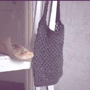 May include: A rustic scene featuring a loaf of bread in a paper bag on a distressed white chair. A dark gray crocheted tote bag hangs from the chair. The background is a textured, light-colored wall.