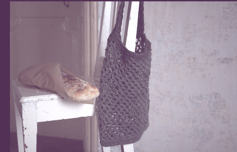 May include: A rustic scene featuring a loaf of bread in a paper bag on a distressed white chair. A dark gray crocheted tote bag hangs from the chair. The background is a textured, light-colored wall.