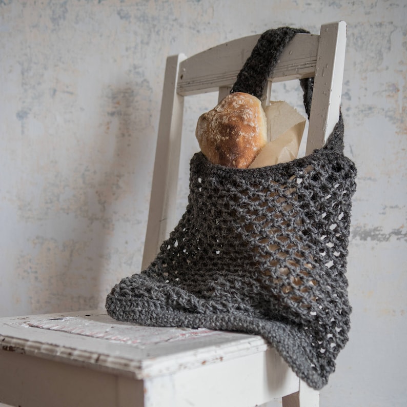 May include: A gray crocheted tote bag filled with a loaf of bread and a paper bag, resting on a white wooden chair. The bag has a net-like design and a long strap. The bread is golden brown.