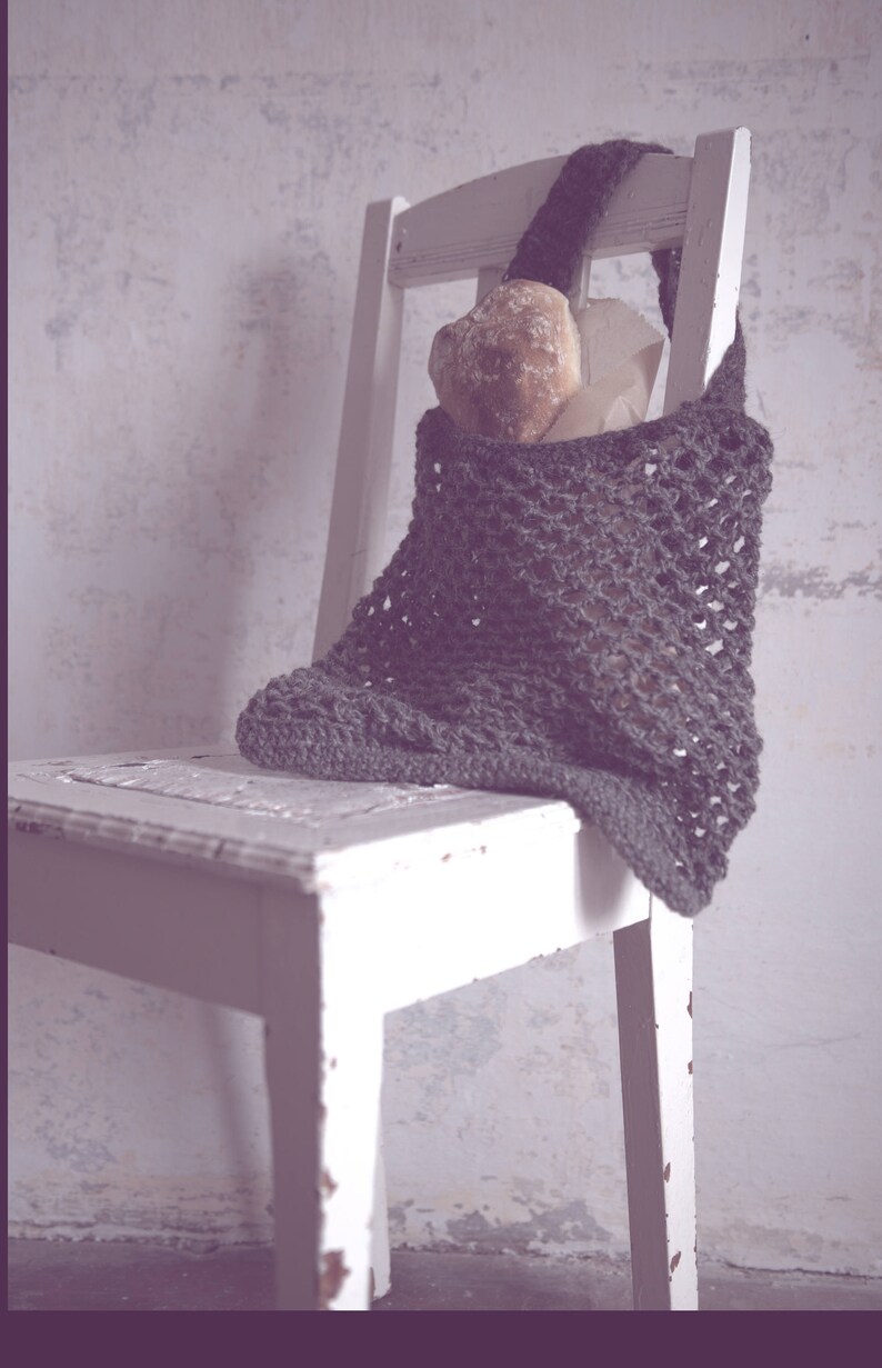 May include: A dark gray crocheted tote bag filled with bread and paper, resting on a white wooden chair. The bag has a large, open weave design and a sturdy handle. The chair has a distressed, vintage look.