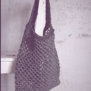 May include: A dark gray, crocheted tote bag hangs from a white wooden chair. The bag has a textured, open weave design. The background is a textured, off-white wall. The bag is a neutral color, suitable for various uses.