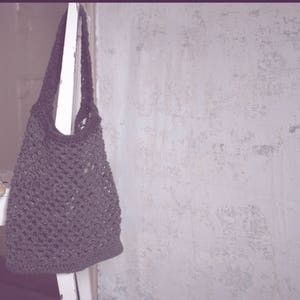 May include: A dark gray crocheted tote bag hangs against a textured white wall. A loaf of bread in a paper bag rests on a small white table. The bag is made of a woven material and has a long strap.