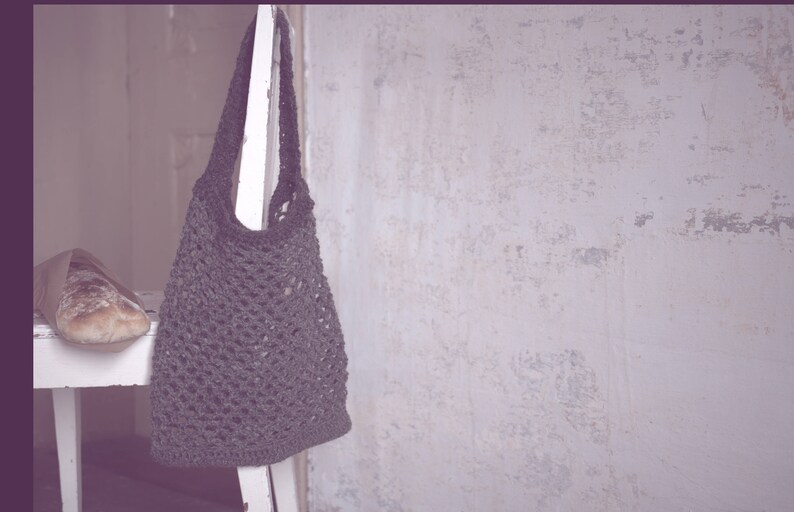 May include: A dark gray crocheted tote bag hangs against a textured white wall. A loaf of bread in a paper bag rests on a small white table. The bag is made of a woven material and has a long strap.
