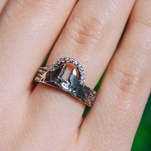 May include: A silver ring with a cutout design of two buildings and a row of small diamonds. The ring is worn on a finger.