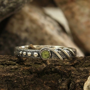 May include: A silver ring with a green gemstone and a textured band. The ring is set on a piece of driftwood.