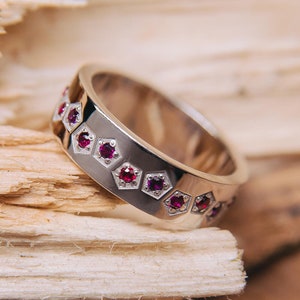 May include: A silver wedding band with a row of small, pink gemstones set in a hexagonal pattern.