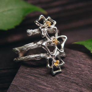 May include: A silver ring with a delicate design of three flowers with yellow gemstones. The ring is set against a dark wooden background with green leaves.