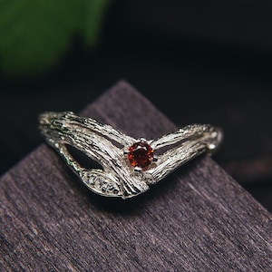 May include: A silver ring with a red gemstone set in a twig-like design. The ring is on a dark wooden surface.