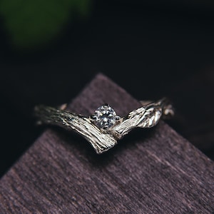 Peut inclure: Une bague de fiançailles en argent avec un seul diamant serti dans une bande en forme de brindille. La bague est présentée sur une surface en bois foncé.