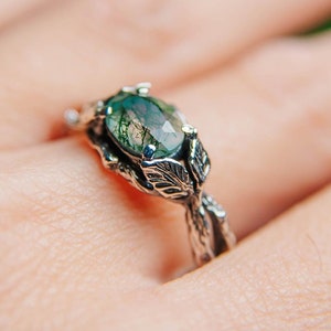 May include: A silver ring with an oval, green gemstone. The ring features a nature-inspired design with leaf and vine details. The gemstone is the focal point, set within a detailed, textured band. The ring is on a hand, with a blurred green background.