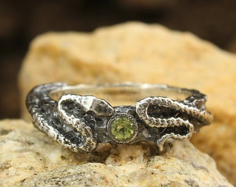 May include: A silver ring with a green gemstone set in a snake design. The ring is on a textured rock surface.