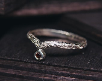 May include: A silver ring with a branch design and a small red gemstone. The ring is on a dark wooden surface.