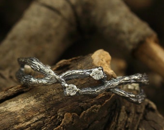 May include: A silver wedding band featuring a delicate, intertwined branch design with small diamonds. The ring is resting on a piece of weathered wood.