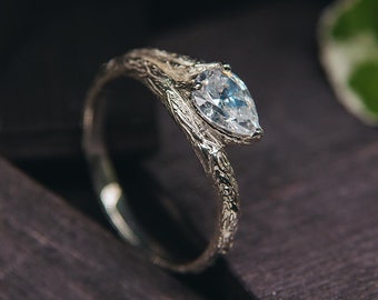 May include: A pear-shaped diamond engagement ring set in a silver band with a unique, branch-like design. The ring is displayed on a dark wooden surface.