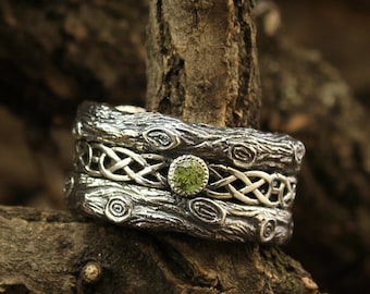 Könnte beinhalten: Ein silberner Ring mit strukturiertem Rindenmuster und keltischen Knoten. Der Ring hat einen kleinen, runden, grünen Edelstein. Der Ring wird auf einem Ast präsentiert, mit dem Logo Wedding Rings Store unten rechts.