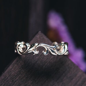 Può includere: Un delicato anello d'argento con un design floreale. L'anello è su una superficie di legno scuro.