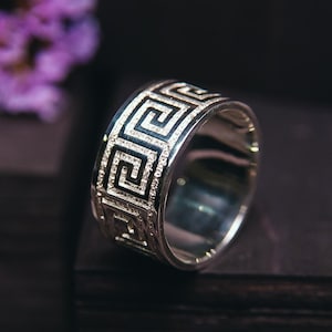 May include: A silver wedding band with a Greek key design. The band is on a dark wooden surface.