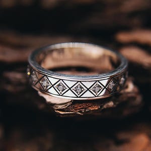 May include: Silver ring with a geometric diamond pattern and small clear stones. The ring is set against a dark, textured background, showcasing its intricate design and craftsmanship. The ring is a wedding ring.