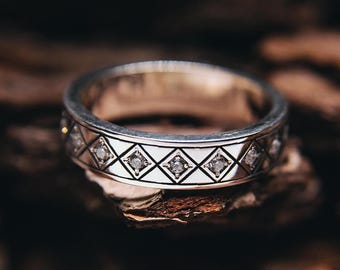May include: Silver ring with a geometric diamond pattern and small clear stones. The ring is set against a dark, textured background, showcasing its intricate design and craftsmanship. The ring is a wedding ring.