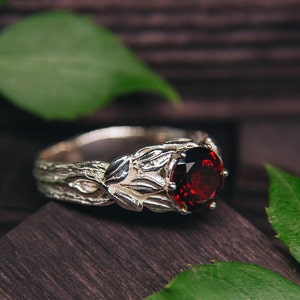 Könnte beinhalten: Ein silberner Ring mit einem roten Edelstein, der in einem Blattdesign gefasst ist. Der Ring liegt auf einer dunklen Holzoberfläche mit grünen Blättern.