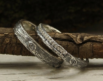 May include: Two silver wedding bands, one with a textured bark-like design and the other with a row of small diamonds. The bands are resting on a piece of weathered wood.