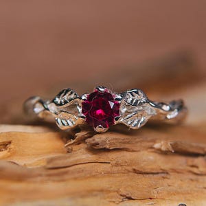 May include: A silver ring with a ruby gemstone set in a leaf design. The ring is on a piece of wood.