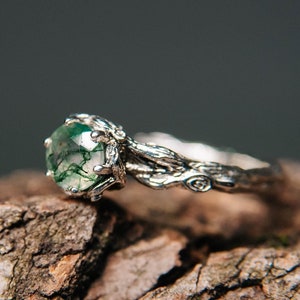 May include: A silver ring with a green moss agate gemstone set in a branch-like band. The ring is resting on a piece of tree bark.