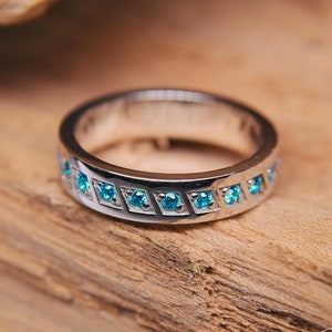 May include: A silver ring featuring a row of small, square, light blue gemstones set in a repeating pattern. The ring is presented on a piece of weathered wood. The Wedding Rings Store logo is visible.