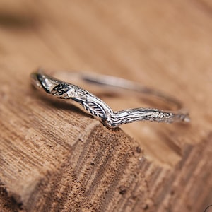 May include: A silver ring with a nature-inspired design. The ring has a V-shaped band with a textured, branch-like appearance and leaf details. The ring is displayed on a wooden surface.