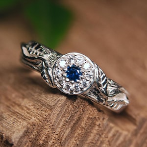 Op de afbeelding: Een zilveren ring met een centrale cluster van kleine witte stenen rondom een diepblauwe edelsteen. De band van de ring heeft een gedetailleerd, door de natuur geïnspireerd ontwerp met blad- en takmotieven.