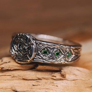 May include: A silver wedding band with a textured design and four green gemstones. The band is on a wooden surface.