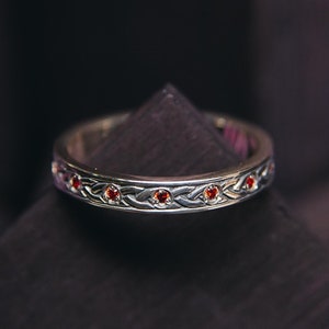 Könnte beinhalten: Ein silberner Ehering mit einem geflochtenen Design und roten Edelsteinen.