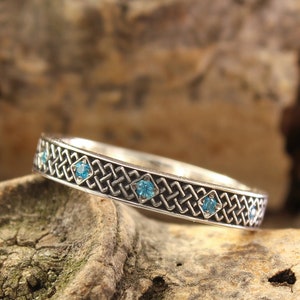 Może przedstawiać: Srebrna obrączka ślubna z celtyckim wzorem węzła i niebieskimi kamieniami szlachetnymi. Obrączka leży na kawałku zwietrzałego drewna.