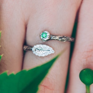May include: A silver bypass ring featuring a textured band resembling a tree branch. One end of the ring is topped with a small, round, vibrant green gemstone, while the other end features a detailed silver leaf shape with a textured surface.