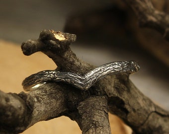 May include: A silver and black wedding band with a textured finish, resting on a piece of wood. The band is shaped like a V.