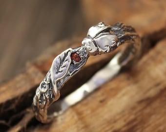 May include: A silver ring with a delicate design featuring a leaf, a small red gemstone, and a butterfly. The ring is set on a piece of wood.