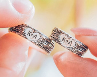 May include: Two silver wedding bands with a tree bark texture. The bands are engraved with a heart and the initials "K+A".