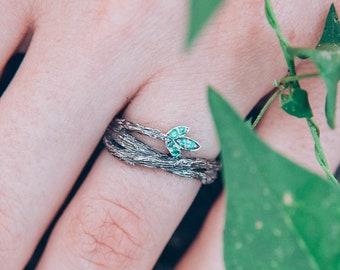 May include: A silver ring with a delicate, twisted band and a small butterfly design with green and blue enamel. The ring is worn on a finger surrounded by green leaves.